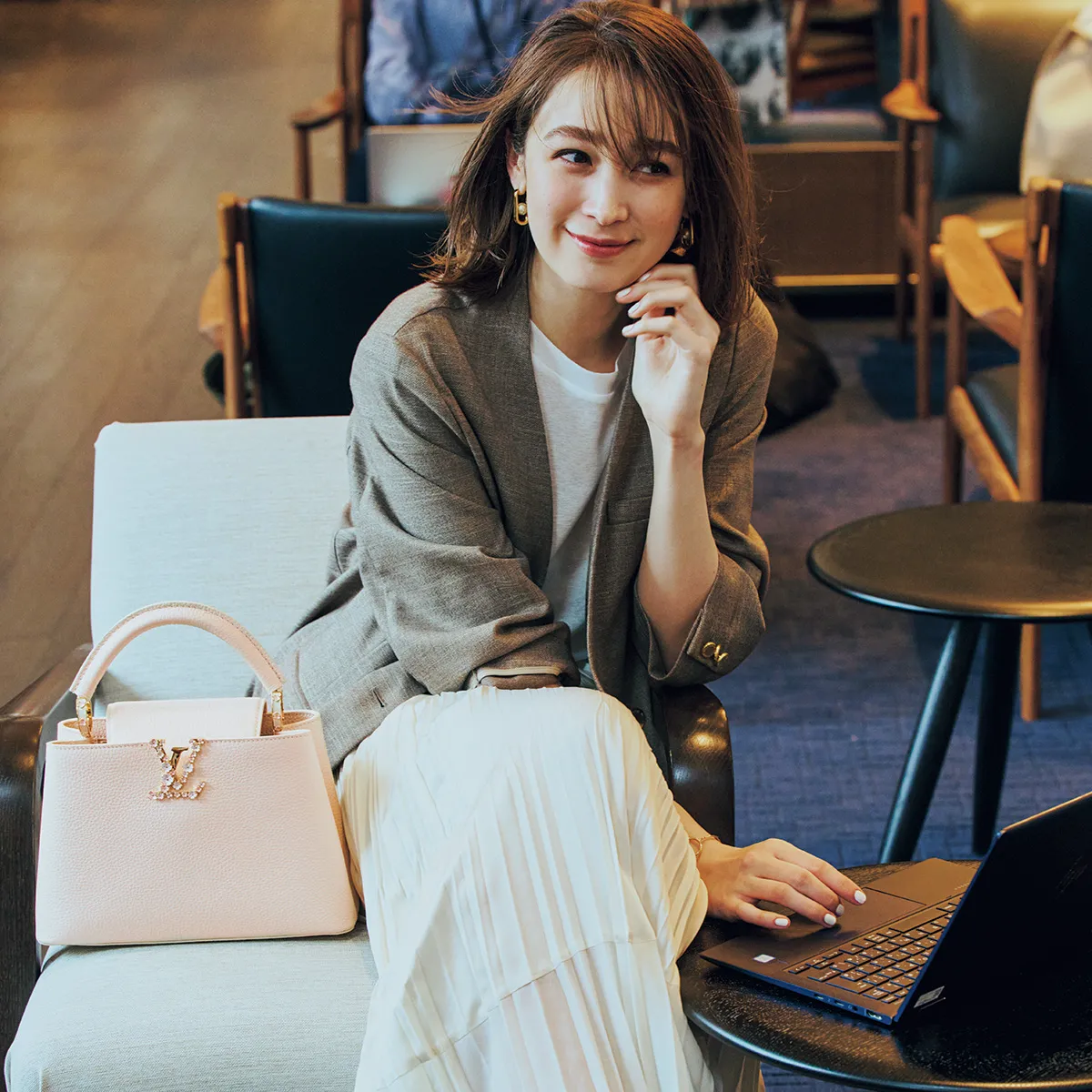 カフェでパソコンを触っている藤井サチさん。ジャケットとプリーツスカートを纏い、耳にはゴールドのフープピアスをつけている。