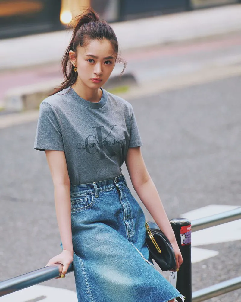グレーのロゴTシャツ、膝丈デニムスカート、レザーのクラッチバッグを合わせた女性