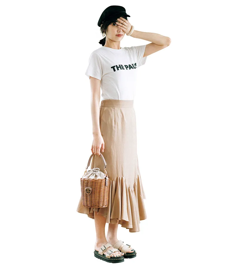 ロゴTシャツ、ベージュのデザインスカート、フラットサンダルを履いた女性