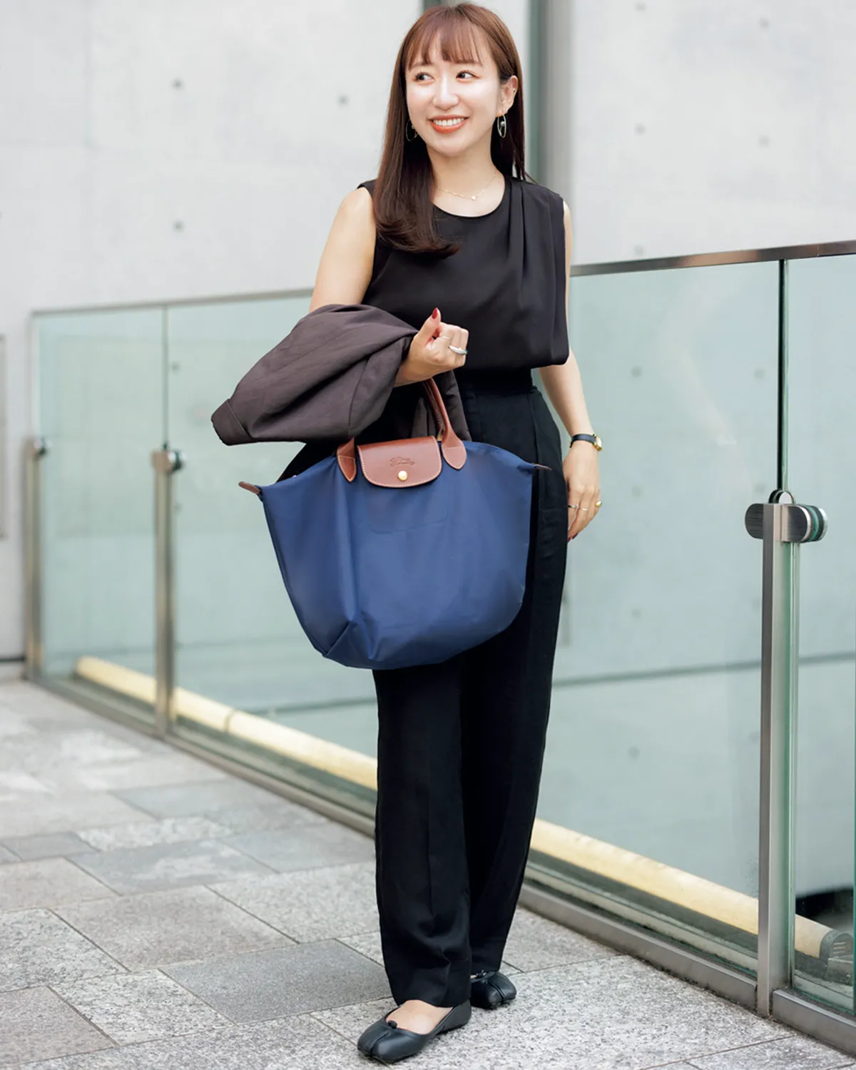 黒のノースリーブトップス×黒パンツの田村有希さん。腕にはグレーのジャケットとネイビーのトートバッグ。